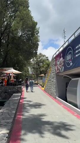 The video shows a pathway in a tourist area. The pathway is paved with gray stones and has a red line along its edge. On the left side of the pathway is a stone wall with greenery behind it. There is a small shop with souvenirs and people around it. A few people walk along the path. On the right is a wall with a large "aba" advertising sign on it, with stairs leading up the wall. The sky is partly cloudy. Tall trees are visible around the path.
