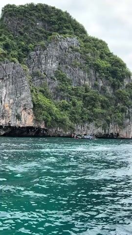 The video shows a body of water with a large, vegetated cliff face in the background. Several boats are visible near the cliff face, and some people appear to be in or around the water. The water is a vibrant turquoise color, and the sky is overcast.