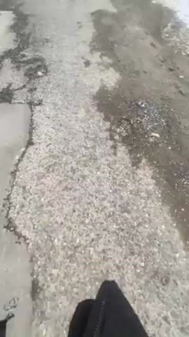 The video shows a walk through a residential area on a cloudy day with some snow on the ground. The camera seems to be held by someone walking, as we see their black pants and shoes at the bottom of the frame. The road is unpaved and appears damaged. There are houses on either side of the road, and utility poles with wires run overhead. The trees lining the street are bare, suggesting it is late fall or winter. The overall atmosphere is bleak and quiet.