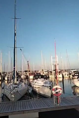 A video captures a serene marina scene in Aabenraa, Denmark. The clear blue sky suggests a pleasant day, likely morning or late afternoon due to the soft lighting. Numerous sailboats and yachts are moored, their masts creating a striking vertical contrast against the horizon. In the foreground, a wooden boardwalk lines the water, with a few life preservers attached to posts. Two sailboats are prominently featured, one a bit larger than the other, docked side-by-side. The water is calm, reflecting the boats and the sky. The overall atmosphere is one of tranquility and peacefulness, typical of a marina on a fine day. There is no visible text in the video.
