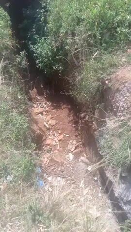 The video shows a narrow, overgrown ditch or trench, possibly a drainage channel, situated in a rural or natural environment. The sides of the ditch are steep and covered with dense green vegetation, including bushes and tall grasses. The ground within the ditch appears to be a mix of reddish-brown soil and scattered rocks and debris, including some white plastic trash. The sunlight suggests it is daytime. The camera is positioned to give a downward view into the ditch as it moves along its length, giving a sense of perspective and depth. The overall impression is one of an unkempt, natural area with signs of human refuse.