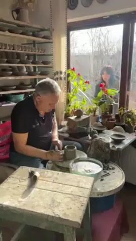 A man is working at a pottery wheel in a bright, cluttered room filled with pottery. He is wearing a black shirt and blue jeans. He sits at the wheel, shaping a small clay pot with his hands. To his left is a small, worn green table with tools on it. Behind him, to the left, are shelves filled with finished pottery pieces. To his right, a window shows a woman standing and watching him from outside. The window sill is adorned with plants and pottery. The room is filled with natural light. A woman's voice speaks in the background.