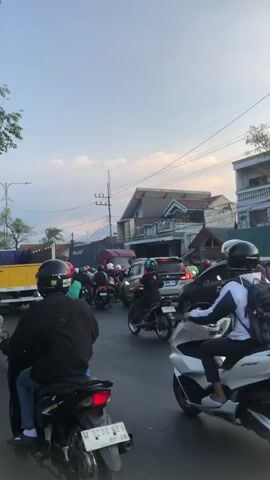 A traffic scene with multiple motorcycles and a few cars on a paved road. The motorcycles are moving in the same direction as the camera, and some are carrying passengers. The environment appears to be a town or city with buildings and overhead wires. The sky is overcast with a hint of sunset colors. One motorcycle in the foreground has its brake lights illuminated. The video is shot from the perspective of someone in or on a vehicle moving with the traffic.