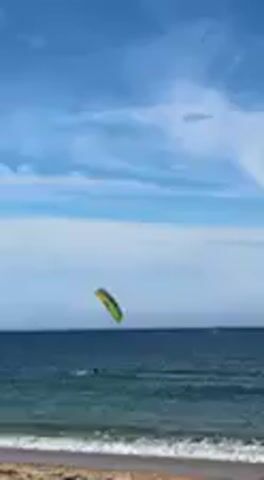 A bright yellow and green kite is visible high in the blue sky with scattered white clouds, over a blue-green ocean. A person is kitesurfing in the distant water, propelled by the kite. Small waves are breaking near the sandy shore. The kite shifts its position in the wind as the kitesurfer navigates the water. The scene takes place during the day, depicting a recreational activity at the beach.