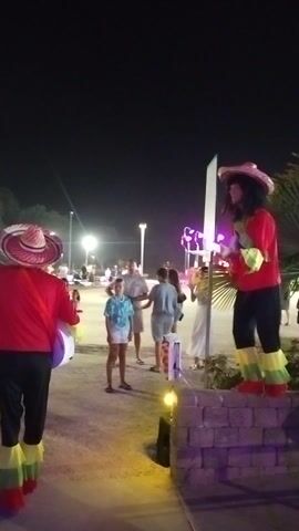 The media file shows a nighttime outdoor scene, possibly a festival or street event in Caorle, Italy. The primary subjects are two costumed figures, dressed in red shirts, black pants, and festive sombreros. Both figures appear to be playing instruments, one with a guitar and the other with a ukulele, suggesting a musical performance. They are also wearing colorful, ruffled boot covers.

Several other people are visible in the background, appearing to be spectators or participants in the event. They are casually dressed and seem to be milling around. The setting includes lighting such as street lamps and decorative, brightly colored lights that illuminate the area. There's also a stone wall visible, possibly part of a building or decorative element. The overall atmosphere suggests a lively and cheerful event. The time of day is clearly night due to the darkness and artificial lighting. There are no specific weather indicators, but the presence of people outdoors suggests it's likely pleasant. No visible text is present in the media file.