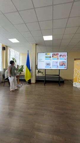 The media file shows a presentation taking place indoors, likely in a conference room or classroom. A woman, dressed in white pants and a grey top, stands on the left side of the frame, holding papers and gesturing towards a large screen. She appears to be speaking to an audience, though only one other person is visible, seated in the background.

The screen displays a presentation with text and images. The Ukrainian flag stands prominently to the left of the screen, suggesting the location is in Ukraine. The room has a wooden floor and a tiled ceiling with fluorescent lighting. There are also plants and chairs visible in the scene. A banner with yellow and white graphics is present on the right side of the screen.

The presentation's title, visible on the screen, appears to be "Напрями роботи і спадщина" which translates to "Areas of Work and Heritage". The images on the screen suggest topics related to community, education, and possibly historical preservation. The overall atmosphere seems professional and informative. The lighting indicates it is daytime, and the indoor setting suggests it could be any weather condition outside.