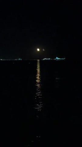 The video is a still image of a dark night scene over water. A bright full moon is visible in the upper center of the frame, casting a shimmering reflection on the water's surface. In the distance, on the horizon, are several illuminated ships or structures. The water itself is dark and appears calm, with the moon's reflection being the primary light source. The overall atmosphere is tranquil and nocturnal.