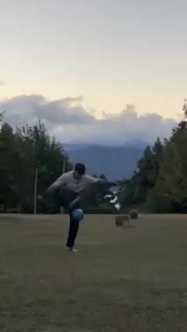 A young man in a gray and black jacket and black pants is juggling a soccer ball on a grassy field. He is using his feet, knees, and thighs to keep the ball in the air. The background includes trees, mountains, and a cloudy sky. The scene appears to be taken during sunset or early evening due to the soft lighting and the color of the sky.