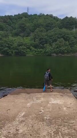 A person stands on a concrete ramp at the edge of a body of water. The person is wearing a backpack and shorts, and holding a blue object. The water is green and appears to be a lake or reservoir. Behind the water, a hill covered in trees rises up. The sky is cloudy. The person appears to be preparing to enter the water or perhaps to fish.