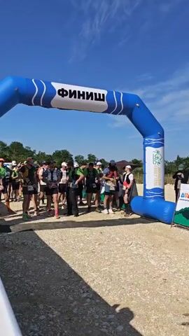 A group of approximately twenty to thirty people, dressed in athletic wear including shorts, t-shirts, and running vests, are gathered under a large blue inflatable arch that says "ФИНИШ" (FINISH) in white letters. Many individuals have hydration packs or small backpacks, and some hold trekking poles. They stand on a dirt and gravel path, facing forward. The scene is outdoors on a sunny day with a clear blue sky and some scattered clouds. Trees are visible in the background. A white barrier runs along the left side of the frame. A male voice speaks in Russian throughout the video.