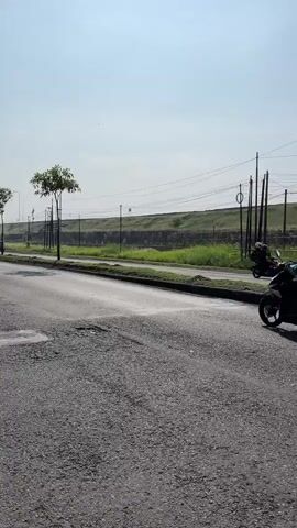 The video shows two people riding motorcycles on a paved road. The road is in disrepair, with visible potholes. In the background, there is a grassy field with a few trees and utility poles. The sky is clear and bright. The person in the foreground is wearing a yellow helmet and a dark jacket. The person in the background is wearing a blue and white jacket. Both are riding away from the camera.