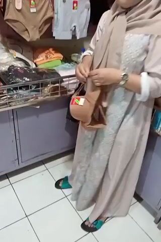 A woman wearing a light brown hijab and a long light brown dress stands in a store. She is holding a small brown purse with a cartoon character on it. The woman is standing in front of a shelf displaying various items of clothing, including underwear, bras, and folded shirts. She appears to be examining or adjusting the purse. The floor is tiled with white and gray tiles.