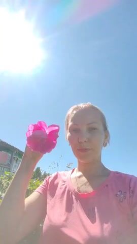 A woman is holding a pink rose to her face, smelling it. She is wearing a pink t-shirt and her hair is pulled back. She has a necklace on. The sun is shining brightly in the blue sky, with lens flare visible. In the background, there is a house and some greenery. The scene appears to be outdoors on a sunny day, possibly in a rural setting like a village. The woman's expression suggests a moment of enjoyment or peace. There is no visible text in the media.