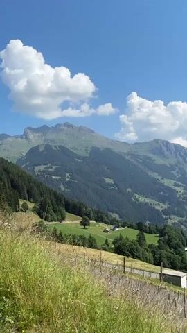 The video is a vertical, panning shot of a mountainous landscape on a sunny day with blue skies and white clouds. The foreground features tall grass and a dirt path or road with a wire fence. In the middle ground, there are rolling green hills dotted with trees and some small buildings, including a rustic wooden house. The background is dominated by large, imposing mountains, some of which appear to have snow-capped peaks. The camera pans from left to right, revealing more of the valley and the distant mountain range.