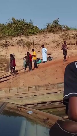 The video shows several people standing on a red dirt bank next to a river or body of water. The camera appears to be located on a boat. Some of the people are dressed in colorful clothing, while one person is wearing a white robe. There is vegetation on the bank behind them. Several of the people are moving around and talking. There is also a structure made of wood on the bank, possibly a makeshift dock or walkway.