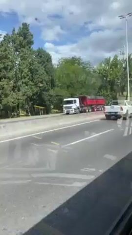 The video is filmed from inside a moving bus looking out the window.  Outside, on a highway, there is a large red dump truck stopped at the side of the road, beyond the guardrail. The bus passes the truck, then passes a white box truck with the word “URBANO” on the side, and the camera view shifts to the inside of the bus.  Several passengers are visible sitting in the yellow seats.