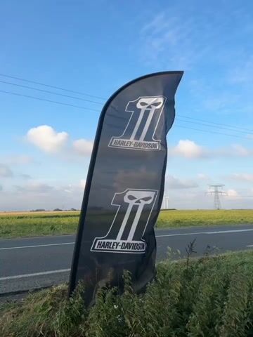 A black flag with the Harley-Davidson logo and the number 1 repeated twice is displayed on a pole. The flag is located outdoors near a road, with a field and power lines visible in the background. The sky is blue with some clouds. The flag is blowing in the wind.