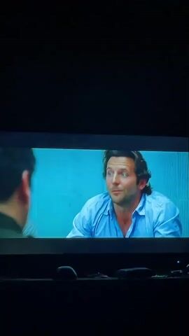 The video shows a screen displaying a scene from a movie or television show. The scene is set in what appears to be an interrogation room with light blue walls. In the initial shot, a man with dark hair and a blue shirt, who appears to be actor Bradley Cooper, is speaking to someone out of frame. The camera angle shifts to show two officers, a man and a woman, both wearing dark green uniforms with gold badges and white shirts. The woman is speaking. The camera then returns to Bradley Cooper, who is now gesturing with his hands as he speaks. The video is being recorded in a dark room, with a couch and other objects visible in the foreground.