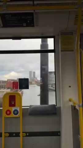 The video shows a view from inside a moving train, looking out the window. Outside, there's a river running through an urban area with buildings of various sizes and designs on either side. The sky is overcast with gray clouds. Inside the train, you can see a yellow safety pole with buttons, a small gray cushion on the window ledge, and signs and posters affixed to the walls and ceiling. The overall lighting is subdued.