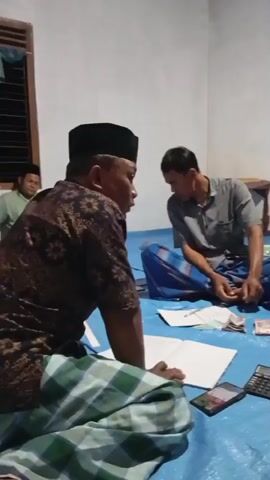 This is a low-resolution video of a group of people sitting on the floor around a blue tarp. Some are wearing traditional Islamic clothing. There are papers, a calculator, and money on the tarp. The people appear to be in a room with white walls. One person is speaking and gesturing with their hands.
