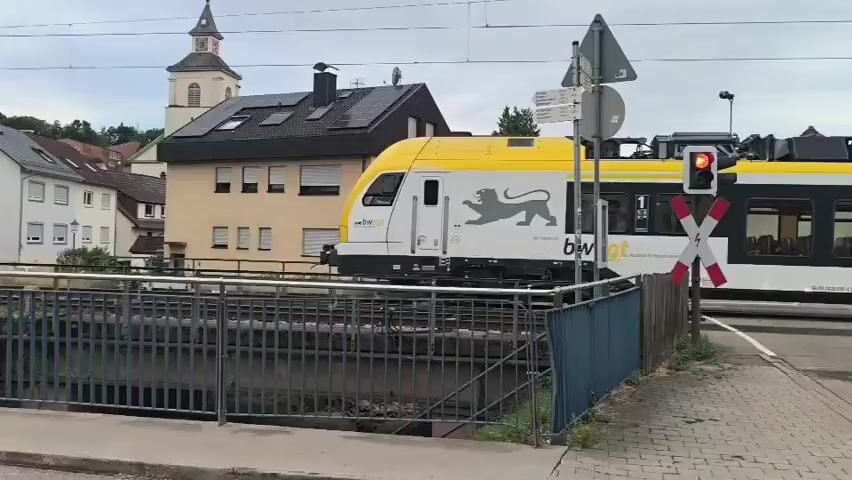A train with yellow and black markings, featuring a lion logo and the text "bw-egt", travels across a bridge over a stream. The train has several windows, some of which show passengers inside. On the left side of the frame, two people on electric scooters are visible. The background shows a town with houses, some with solar panels on their roofs, and a church steeple. The sky is overcast. A railroad crossing signal with flashing lights is also present.