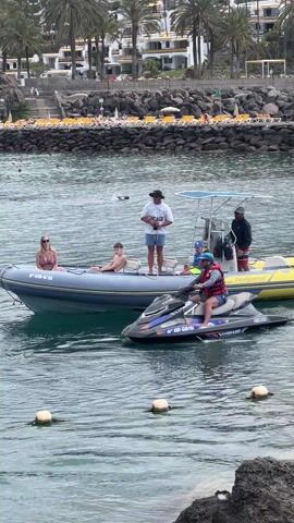 The video shows a beach scene on a sunny day. A group of people are on a grey inflatable boat and another person is on a black and blue jet ski. A man is standing on the boat wearing a white shirt with the word "CHICAGO" written on it, blue shorts, and a black hat. A woman in a bikini and a young boy are sitting on the boat. Another man in a hat and a dark shirt stands near the boat's controls. A young boy in a life vest is on the boat as well. The person on the jet ski is wearing a life vest and shorts. A line of buoys stretches into the water from a rocky outcrop. In the distance, a sandy beach is filled with sunbathers and umbrellas under palm trees. Someone jumps into the water and swims.