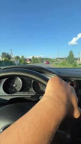 A first-person perspective video shows a person driving a car on a road. The driver's right hand is on the steering wheel. The dashboard shows the speedometer is around 65-68 kilometers per hour. The car is on a paved road with a clear blue sky and some white clouds. There are trees and houses visible on the sides of the road. A red car is ahead on the road.