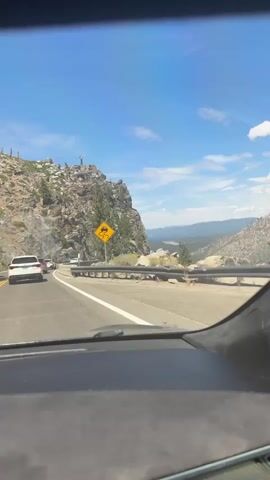 This video captures a view from inside a car driving on a mountainous road on a sunny day. The road curves to the right, bordered by a concrete barrier and a steep, rocky cliff face on the left. Several cars are visible ahead, also traveling along the road. A yellow diamond-shaped sign with a black symbol depicting a car skidding is prominently displayed on the left side of the road, indicating a potential hazard. Pine trees and sparse vegetation dot the rocky landscape. In the distance, rolling hills and a valley can be seen under a clear blue sky with scattered white clouds.