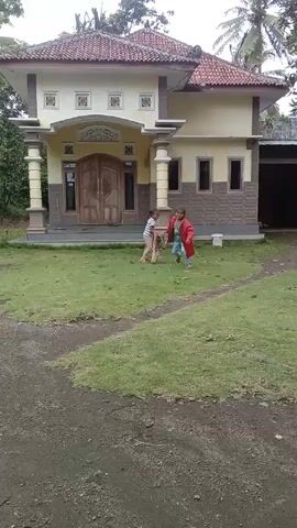 Two young girls are running in the front yard of a house on a cloudy day. The house has a tan exterior with gray stone accents, a red tile roof, and a double-door entrance flanked by columns. The yard is covered in grass and a dirt driveway leads to the house. The girl in the front is wearing a red jacket and jeans and is running toward the camera. The second girl is carrying a brown jacket over her shoulder and smiling as she chases after the first girl. Palm trees and other vegetation are visible in the background.