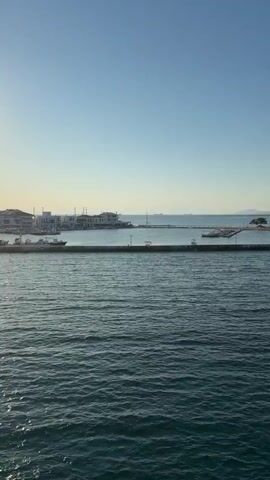 The video shows a wide shot of a body of water with a clear blue sky. In the distance, a white building with a red roof sits on a small island connected to a pier. There are some trees on the island. Along the pier, there are lights and what appear to be benches. Further in the background, there are hills and more structures, possibly part of a town. On the water, there are a few boats, including a small white one near the pier. The water is calm with gentle ripples.