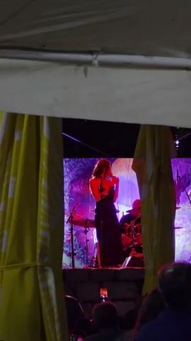 A female singer in a black dress performs on a stage, illuminated by purple and pink lights. She is holding a microphone and standing in front of a drummer. The stage is set against a backdrop that appears to be a projected image of a forest or nature scene. The scene is viewed through an opening framed by yellow and white fabric, possibly part of a tent or awning. Several audience members are visible in the foreground, their faces partially obscured.