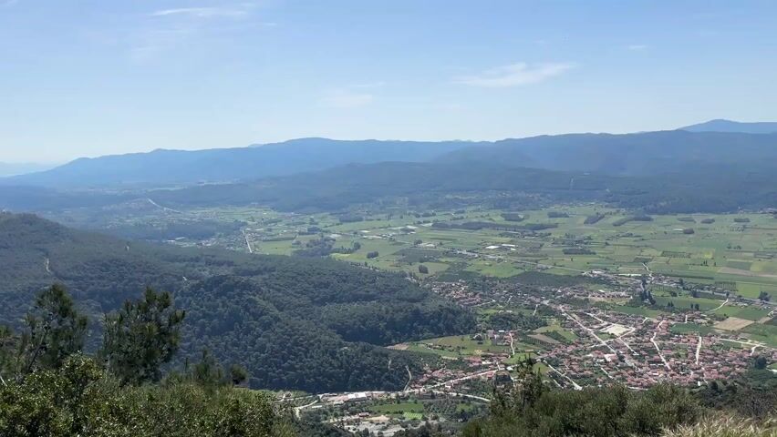 The video shows a scenic view of a valley from a high vantage point. The landscape includes a mix of dense forests, agricultural fields, and a small town with red-tiled roofs. In the background, there are mountains under a clear blue sky. The camera pans slightly to the right, revealing more of the valley and a tree on the right edge of the frame. There are no people visible in the scene. The overall impression is a peaceful, rural landscape.