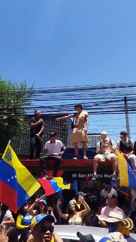 The video shows a lively outdoor scene with a crowd of people, many holding and waving Venezuelan flags. Several individuals are sitting and standing on top of a vehicle, possibly a bus, that has signage indicating "San Alberto Hurtado - Padre Manuel Daza." The people on top are waving flags and appear to be celebrating or protesting. One man, shirtless with a white headband, waves a flag enthusiastically. The sky is clear and blue, and power lines are visible above. The general atmosphere is festive and spirited.