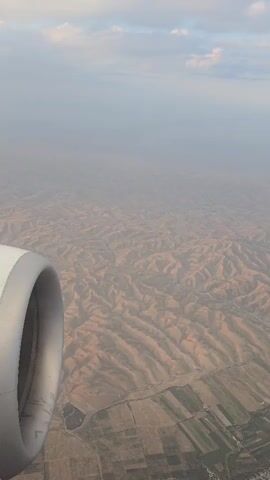 The video shows an aerial view from an airplane window. In the foreground, the engine cowling of the aircraft is visible on the left side. The background shows a vast, arid landscape with rolling hills and valleys. The terrain appears dry and sparsely vegetated, with some patches of green, cultivated fields visible in the distance. The sky is hazy with scattered clouds. The camera is relatively stationary, offering a static perspective of the landscape below.