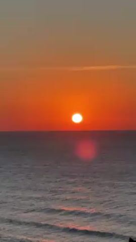 The video shows a sunset over the ocean. The sun is a bright orange orb, with the sky a similar color. The ocean is a calm gray. There are small waves on the surface of the water. There is a lens flare in the center of the view. A boat is visible in the distance.