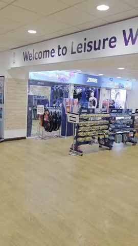 The video is taken inside a Leisure World building, a multi-purpose leisure facility. The camera first focuses on the entrance to the Leisure World store, which sells swimwear and accessories. The camera then pans towards the reception area for the Charter Hall. The area is relatively empty, with only a few people walking in the distance. Finally, the camera focuses on a poster advertising the Rapture Gaming Festival, held at the Charter Hall in Colchester. The poster features a variety of characters and states the festival dates as August 1st and 2nd, 2026.