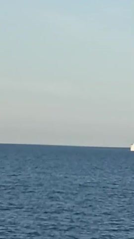 A long shot video captured on a clear day shows a vast expanse of blue ocean under a pale blue sky. A small white catamaran is visible on the left side of the frame, far in the distance. As the video progresses, a large red and white cargo ship, identified by the name "MURASTA" on its side, moves from left to right across the frame. The cargo ship is significantly larger than the catamaran and is the primary focus of the video. The movement of the cargo ship suggests it is navigating through the open sea.