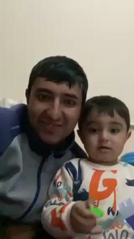 A medium shot shows an adult man and a young child sitting together looking at the camera. The man is dark-haired, light-skinned, and smiling. He wears a grey and dark blue jacket. The child is also dark-haired and light-skinned, wearing a patterned long-sleeved shirt. The child holds a small green toy. The background is a plain white wall.