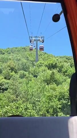 The video shows a view from inside a cable car moving uphill. Several orange and white cable car cabins are visible on the wires, some moving towards the camera and others moving away. The cable cars are suspended over a lush, green, tree-covered mountainside under a clear blue sky. The interior of the cable car is partially visible, including a gray seat in the foreground and an orange frame around the window.