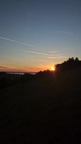 The video shows a sunset over a hilly landscape. The sky is a gradient of blue and orange, with contrails from airplanes visible. The sun is setting behind a silhouetted treeline on a hill. In the distance, there is a body of water and a town. The foreground is a dark, grassy hillside.