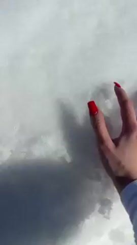 The video begins with a close-up shot of a hand with red nail polish touching fresh snow. The camera then pans out to show a snow-covered landscape with tire tracks. A person dressed in a white outfit is seen running across the snowy field, kicking up snow as they move. The scene is bright and sunny, with a clear blue sky and trees visible in the background. The environment appears to be a rural or open area covered in snow, possibly a field or a hillside.