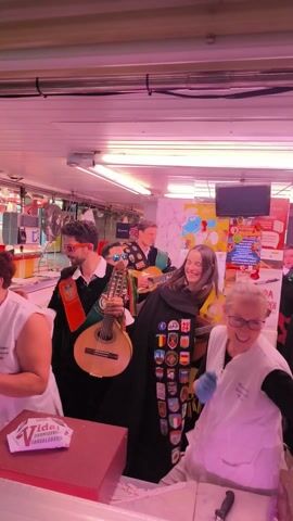 Inside a brightly lit fish market, a musical troupe is performing for the staff. The troupe consists of at least four individuals, dressed in costumes that include capes adorned with patches, and playing instruments like a mandolin and guitars. Two female fishmongers, wearing white uniforms and blue gloves, are visible behind the fish counter, smiling and engaging with the performance. The market setting is evident from the display cases filled with seafood and the scales used for weighing purchases. The atmosphere appears jovial and celebratory, with the music and interactions suggesting a special occasion.