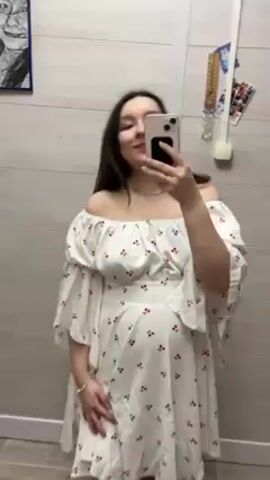 A woman with long dark hair is taking a selfie in front of a mirror. She is wearing a white, off-the-shoulder dress with a red cherry print. The dress has flowy sleeves and a slightly cinched waist, emphasizing a noticeable baby bump. She is also wearing a simple necklace and bracelet. The background consists of a tiled wall, a partial view of a light wood-colored cabinet, and a few miscellaneous items hanging on the wall. The woman smiles slightly and makes a small hair flip gesture. The camera, likely her smartphone, is held up in front of her face, partially obscuring it.