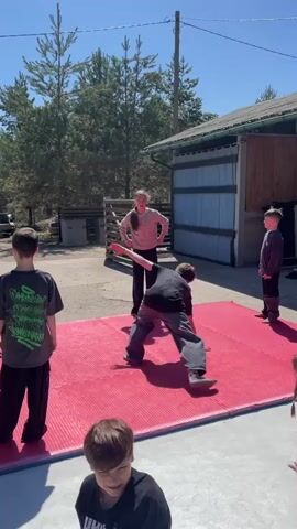 A group of children are outside learning breakdancing moves from an older girl standing among them. They are on a red mat in a yard, near a brown wood and metal garage-type building.  Beyond the yard, a swimming pool is seen with a bright blue cover. The pool is bordered by deck and lawn, and surrounded by tall, bare birch trees. The sky is bright blue with very few clouds.  Some of the children are focused on copying the breakdance moves.