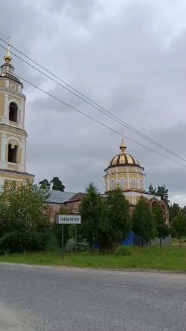 A video shows a church tower and a domed church. A sign in front of the church reads "ПАШНЕВО". The sky is overcast and cloudy. Trees and bushes surround the church. A road is visible in the foreground.