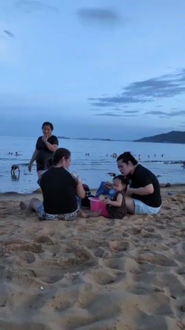 The video shows a beach scene at dusk. Three women and a young child are sitting on the sand in the foreground, appearing to be playing or interacting. Two women are seated with their backs to the camera, and one is looking towards the child. The child is sitting between them, holding a pink bucket and some toys. A third woman is standing further back, closer to the water, and seems to be looking towards the sea. In the background, several people are visible in the shallow water, and a landmass with hills is visible in the distance. The sky is a pale blue with some clouds, indicating late afternoon or early evening. The sand is light brown and textured with footprints and ripples.
