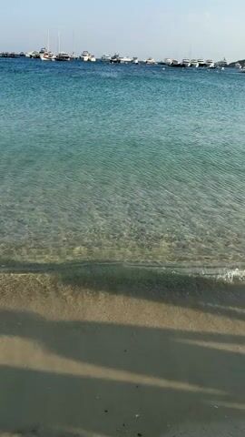 The video captures a tranquil scene at Agios Ioannis Diakoftis, Greece. The foreground is dominated by a sandy beach with the gentle lapping of clear, shallow turquoise water. Sunlight casts dappled shadows across the sand, suggesting it's either morning or late afternoon.

The middle ground features the shimmering expanse of the Aegean Sea, so clear that the sandy seabed is visible beneath the surface. A few individuals are casually swimming or wading in the water, enjoying the calm conditions.

In the background, a picturesque coastline unfolds. White, cubist-style buildings are scattered across a hillside, characteristic of Greek island architecture. A line of boats, including sailboats and yachts, are anchored in the bay, adding to the serene maritime atmosphere. The sky is a clear, vibrant blue, indicating pleasant weather. The overall mood is peaceful and idyllic, showcasing a beautiful Mediterranean beach day. No visible text is present in the media.