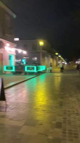 The video shows an outdoor area at night, possibly a bar or restaurant. There are people seated at tables and a live band performing on a small stage. The area is lit by neon lights and string lights. There are signs visible, one of which says "WE/ON BAR". A screen displays the text "Alcohol Te está llamando". The ground is wet, suggesting it may have recently rained. The camera is moving, giving a dynamic but somewhat blurry view of the scene.