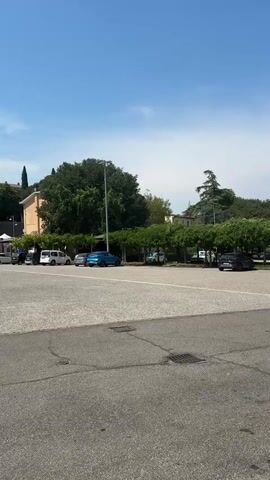 The video shows a wide shot of a town square on a sunny day. The square is paved with light-colored cobblestones and asphalt. Several cars are parked in the square, including a white van, a blue sedan, and a dark-colored SUV. A row of neatly trimmed green hedges runs along one side of the square, separating it from a street. Behind the hedges, there are several buildings with light-colored facades and tiled roofs. Some of these buildings appear to be houses, while others might be businesses. A tall, thin lamppost with two lights stands in the middle of the square. In the background, a hill is covered with trees and more buildings, including a building with a clock tower. The sky is clear and blue with a few wispy clouds. The video pans slowly from right to left, offering a panoramic view of the square and its surroundings. There are no people visible in the video.