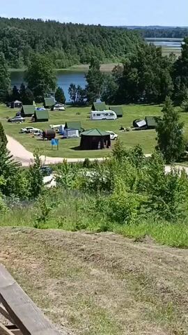 The video shows a panoramic view of a campground on a sunny day. In the foreground, there is a grassy slope with some dried grass and a wooden railing. The middle ground features a campground with several small cabins, tents, and campers. There are also cars parked around the campground. In the background, there is a body of water, likely a lake, surrounded by lush green trees. The sky is blue with some scattered clouds. The overall scene depicts a peaceful outdoor recreational area.